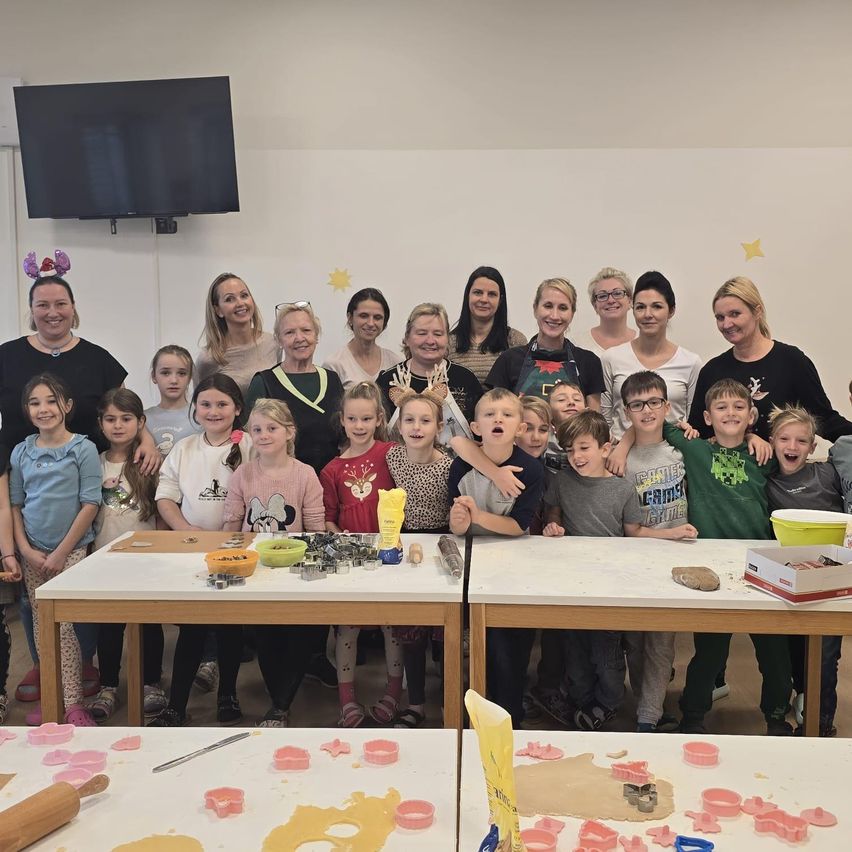 Eine Gruppe von Erwachsenen und Kindern versammelt sich in einem Raum für eine Backaktivität. Sie stehen hinter Tischen mit Teig und Ausstechformen. Eine Frau trägt eine Weihnachtsschleife.