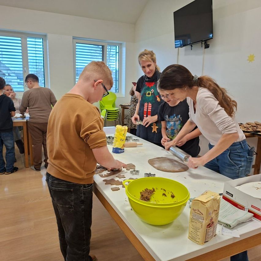 In einem Raum versammeln sich mehrere Personen um einen Tisch und beschäftigen sich mit einer Backaktivität. Ein Junge arbeitet am Teig, während zwei Frauen in der Nähe sind, eine hält eine Nudelrolle. Der Tisch ist mit Backutensilien und Zutaten übersät. Im Hintergrund sitzen weitere Personen an anderen Tischen.
