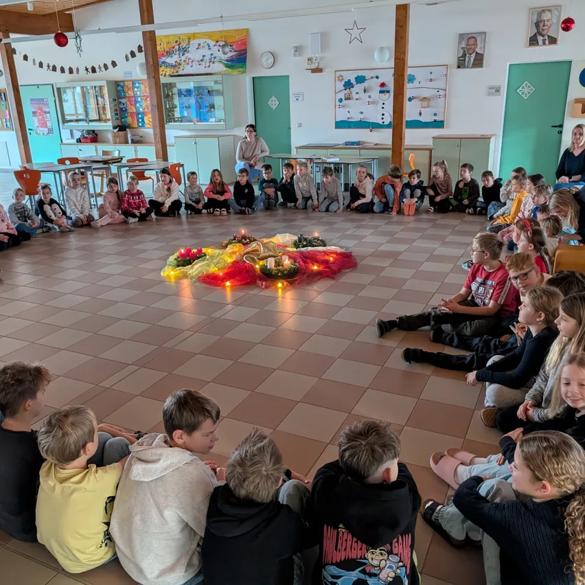 Kinder sitzen in einem Kreis um eine kerzenbeleuchtete Dekoration in einem Klassenzimmer, während Erwachsene von den Seiten beobachten. Der Raum ist mit Feiertagsdekorationen geschmückt.
