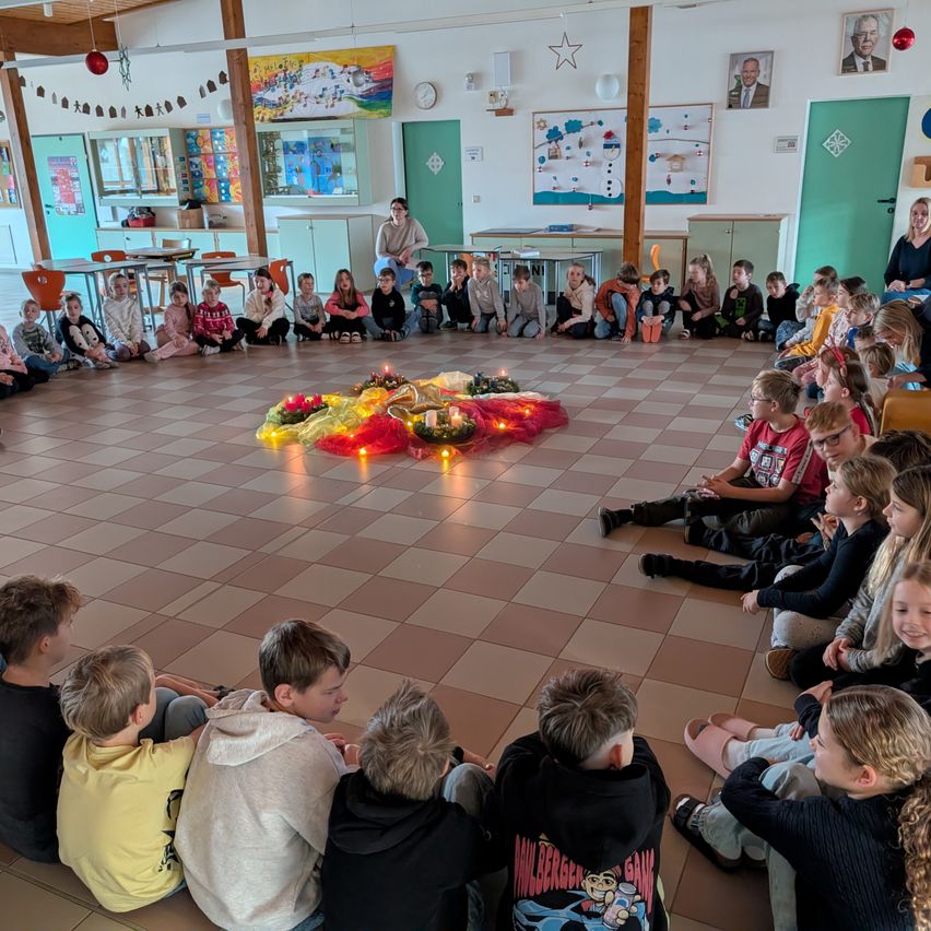 Kinder sitzen in einem Kreis um eine kerzenbeleuchtete Dekoration in einem Klassenzimmer, während Erwachsene von den Seiten beobachten. Der Raum ist mit Feiertagsdekorationen geschmückt.
