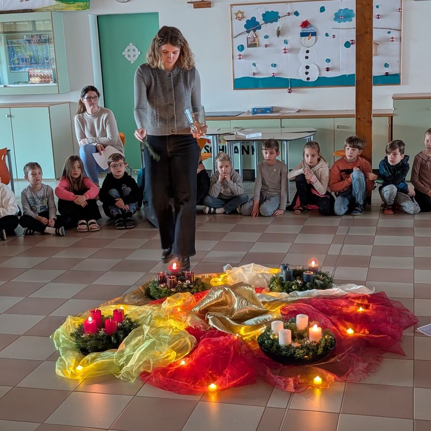 Eine Frau steht in einem Raum mit einer Gruppe von Kindern, die auf dem Boden sitzen, um eine Krone mit brennenden Kerzen und Dekorationen auf dem Boden herum.
