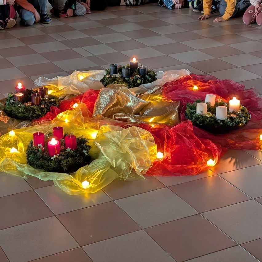 Ein gefliester Boden ist mit Kränzen aus grünen Blättern und roten, gelben und weißen Kerzen geschmückt. Die Kränze sind beleuchtet und werfen ein warmes Licht. Menschen sitzen um die Kränze auf dem Boden.
