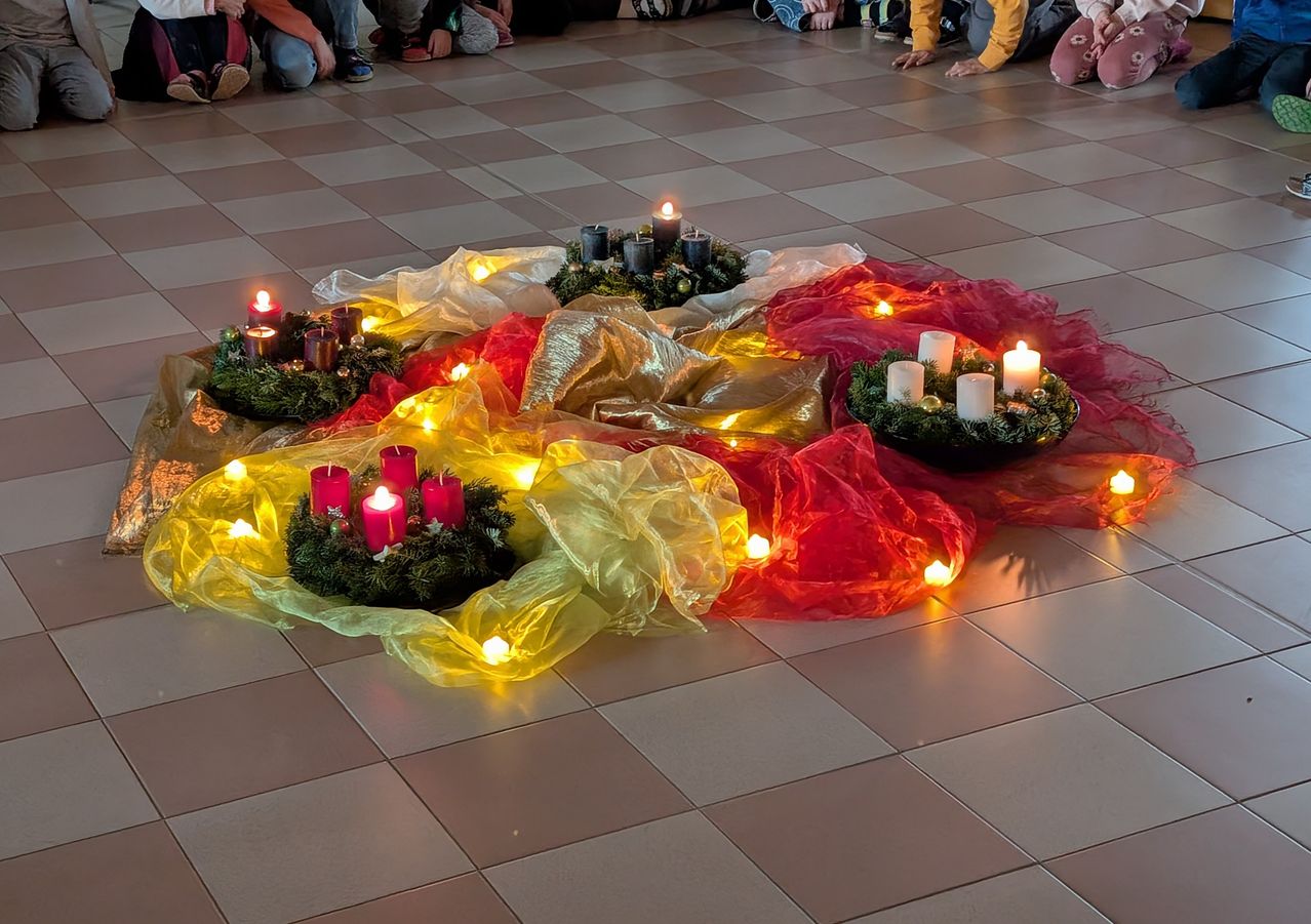 Ein gefliester Boden ist mit Kränzen aus grünen Blättern und roten, gelben und weißen Kerzen geschmückt. Die Kränze sind beleuchtet und werfen ein warmes Licht. Menschen sitzen um die Kränze auf dem Boden.