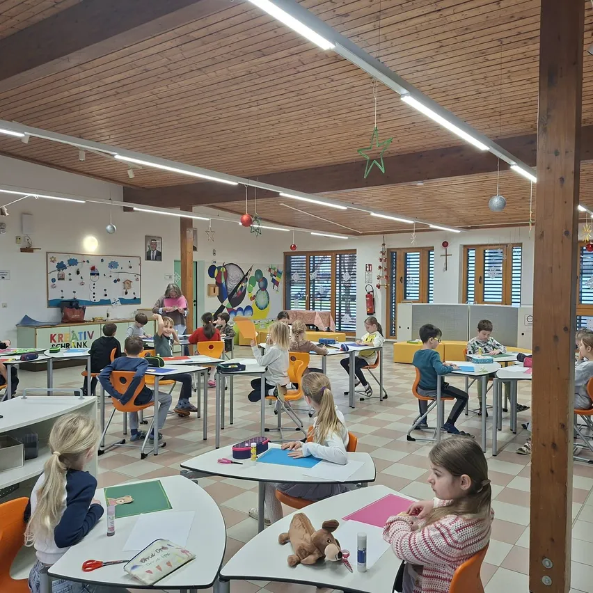 Ein Klassenzimmer mit Kindern an Tischen, einer Lehrerin, die in der Nähe einer Tafel steht, Holzbalken an der Decke und verschiedenen Klassenzimmerdekorationen.