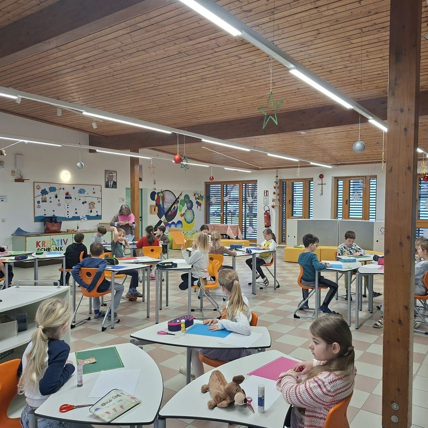 Ein Klassenzimmer mit Kindern an Tischen, einer Lehrerin, die in der Nähe einer Tafel steht, Holzbalken an der Decke und verschiedenen Klassenzimmerdekorationen.