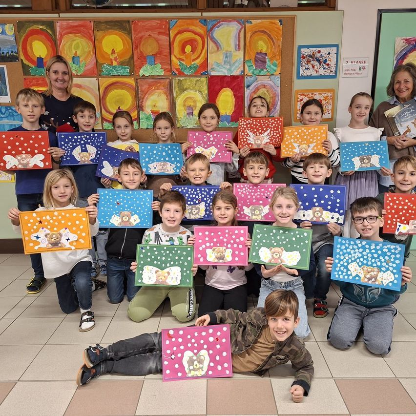 Eine Gruppe von Kindern in einem Klassenzimmer posiert mit ihren farbenfrohen Kunstwerken. Sie halten bemalte Leinwände mit verschiedenen Designs. Der Raum ist mit hellen, festlichen Gemälden an den Wänden dekoriert.