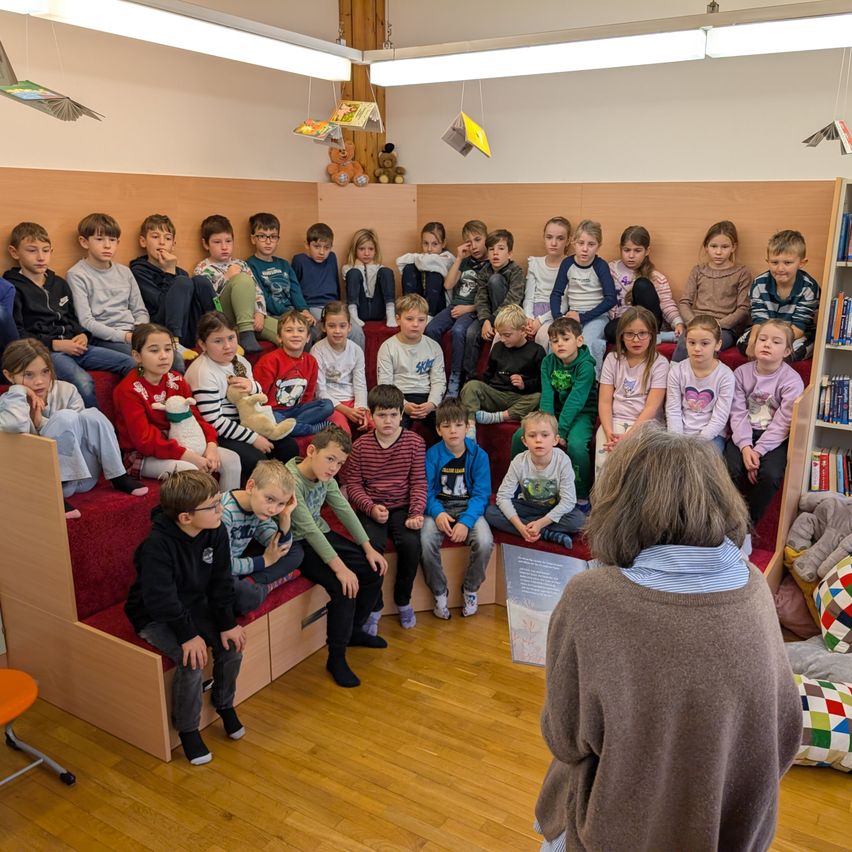 Ein Klassenzimmer mit Holzbänken und Regalen voller Bücher. Kinder sitzen auf den Bänken und hören aufmerksam einer Frau zu, die vor ihnen steht. Einige haben Kuscheltiere.