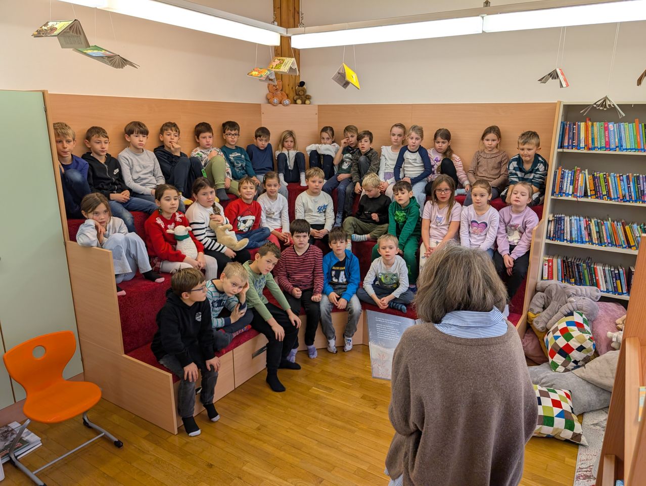 Ein Klassenzimmer mit Holzbänken und Regalen voller Bücher. Kinder sitzen auf den Bänken und hören aufmerksam einer Frau zu, die vor ihnen steht. Einige haben Kuscheltiere.