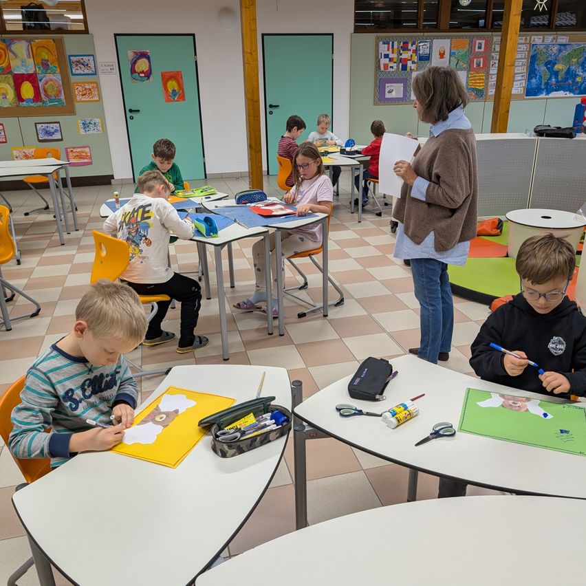 Ein Klassenzimmer mit Schülern, die an Kunstprojekten arbeiten. Eine Frau steht vorne und hält ein Blatt Papier. Kinder sitzen an Tischen mit Kunstmaterialien, und farbenfrohe Kunstwerke schmücken die Wände.