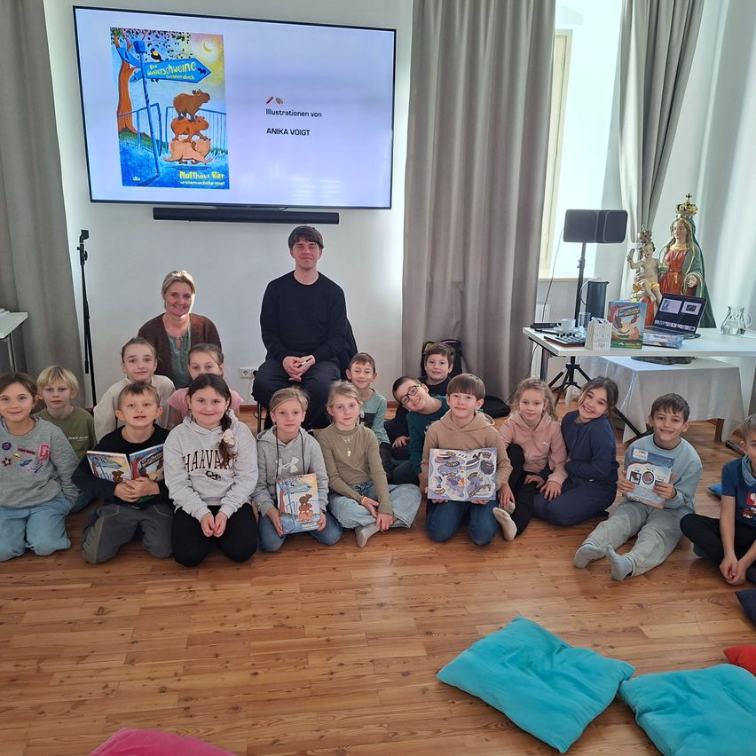 Eine Gruppe von Kindern sitzt auf dem Boden, liest Bücher und schaut zu einem Mann und einer Frau, die vor einer Projektionsleinwand stehen.