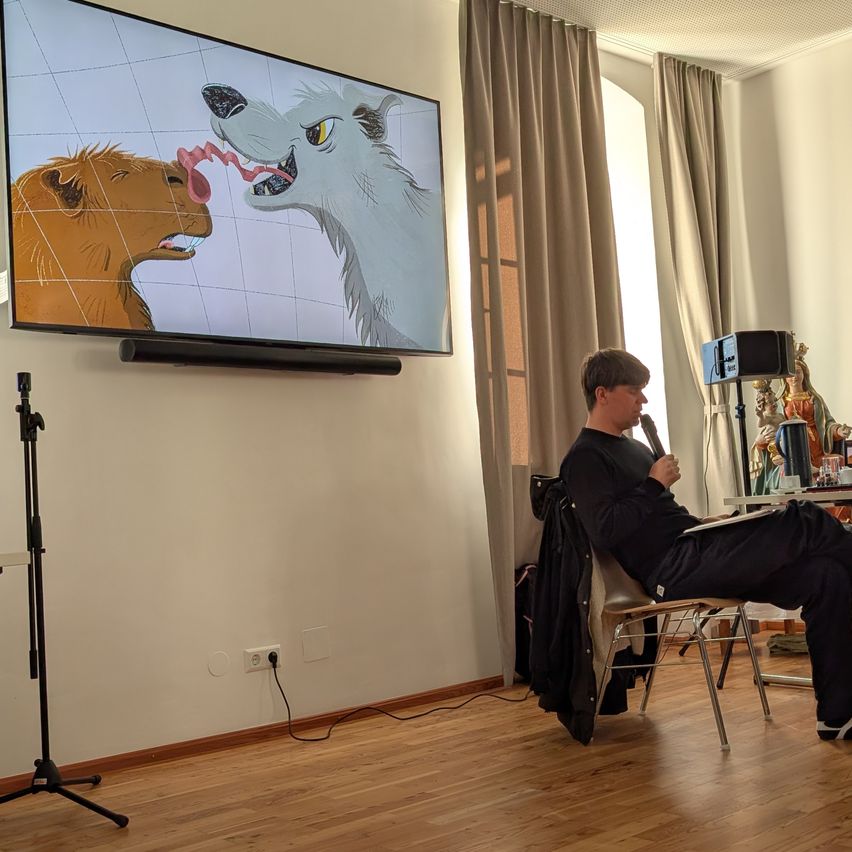 Ein Mann sitzt auf einem Stuhl und hält ein Mikrofon. Ein Fernseher ist an der Wand montiert und zeigt ein Bild von zwei Tieren.