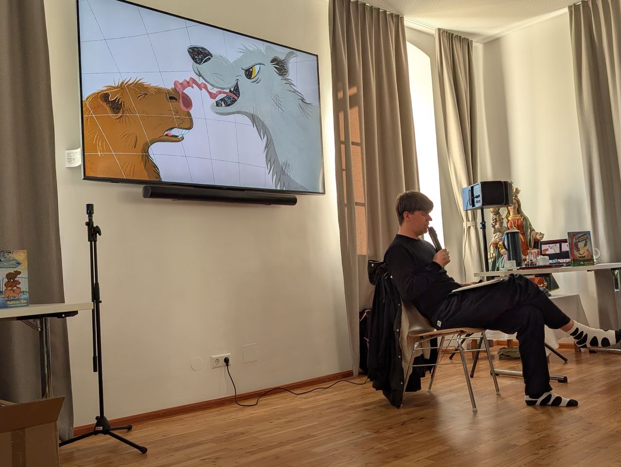 Ein Mann sitzt auf einem Stuhl und hält ein Mikrofon. Ein Fernseher ist an der Wand montiert und zeigt ein Bild von zwei Tieren.