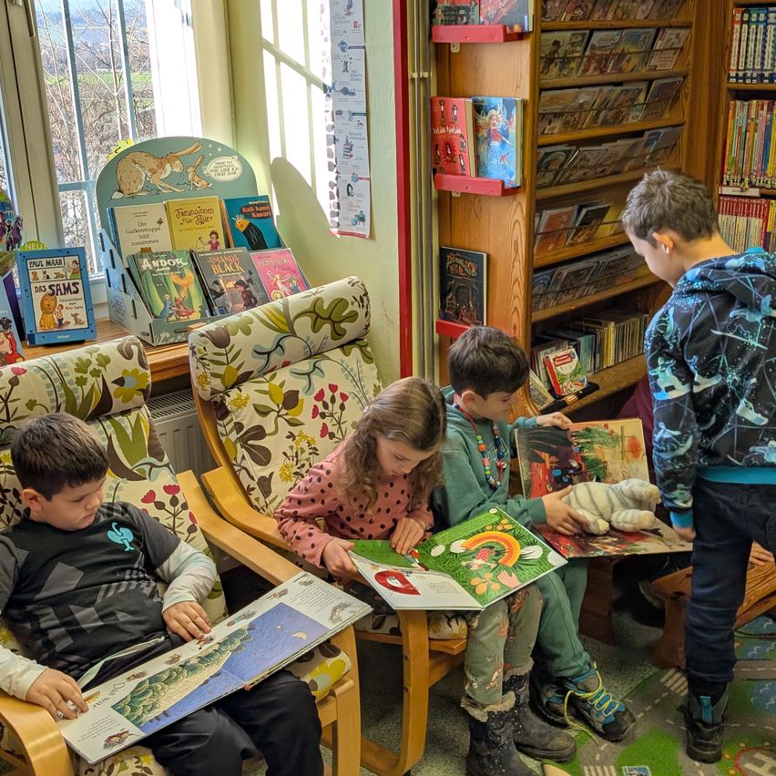 Vier Kinder sitzen in einer Bibliothek, jedes vertieft in sein eigenes Buch. Sie sitzen auf farbenfrohen Stühlen, ein Junge liest links, ein Mädchen im Punkteshirt liest in der Mitte und zwei andere Jungen sitzen rechts. Dahinter ist ein Bücherregal mit Büchern zu sehen.