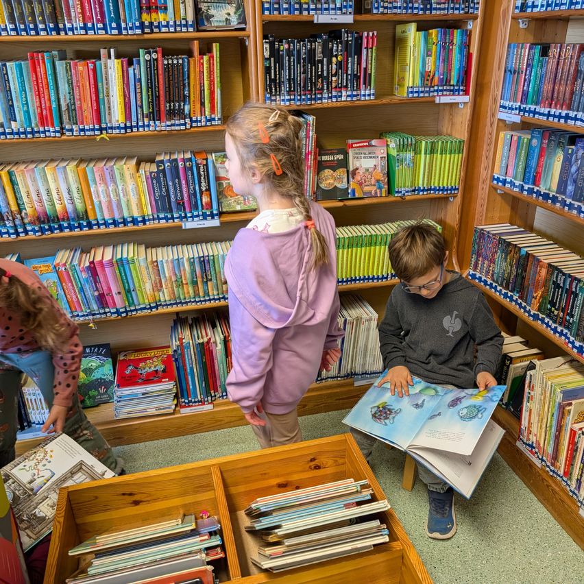 Drei Kinder erkunden eine Bibliothek. Ein Junge liest ein Buch, während ein Mädchen mit Zöpfen neben einem Bücherregal steht. Ein anderes Kind schaut sich Bücher auf einem unteren Regal an.