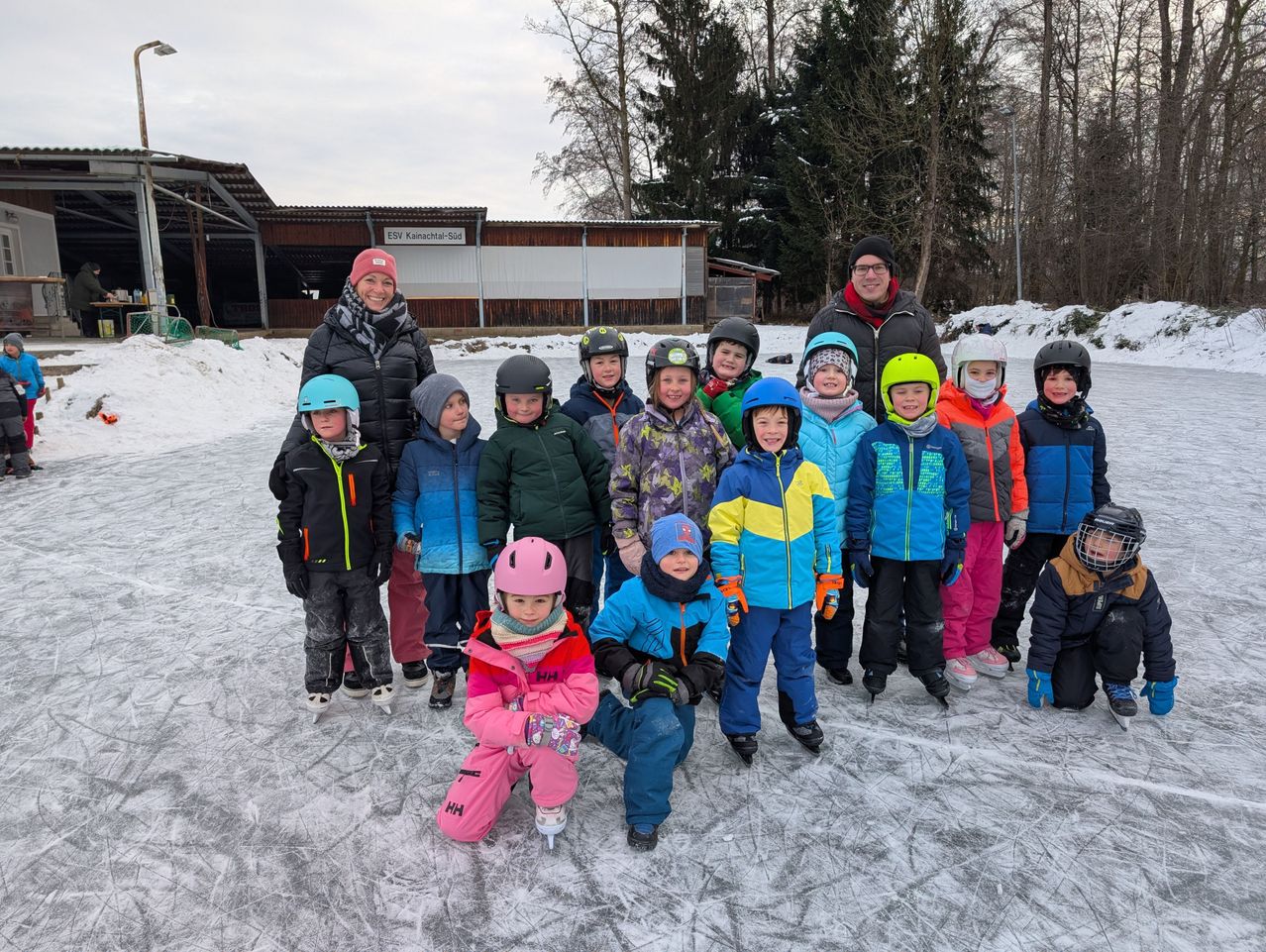 Eine Gruppe von Kindern und Erwachsenen posiert für ein Foto auf einer Eisbahn. Dahinter steht ein Gebäude mit der Aufschrift ESV Kleinmachnow Süd.