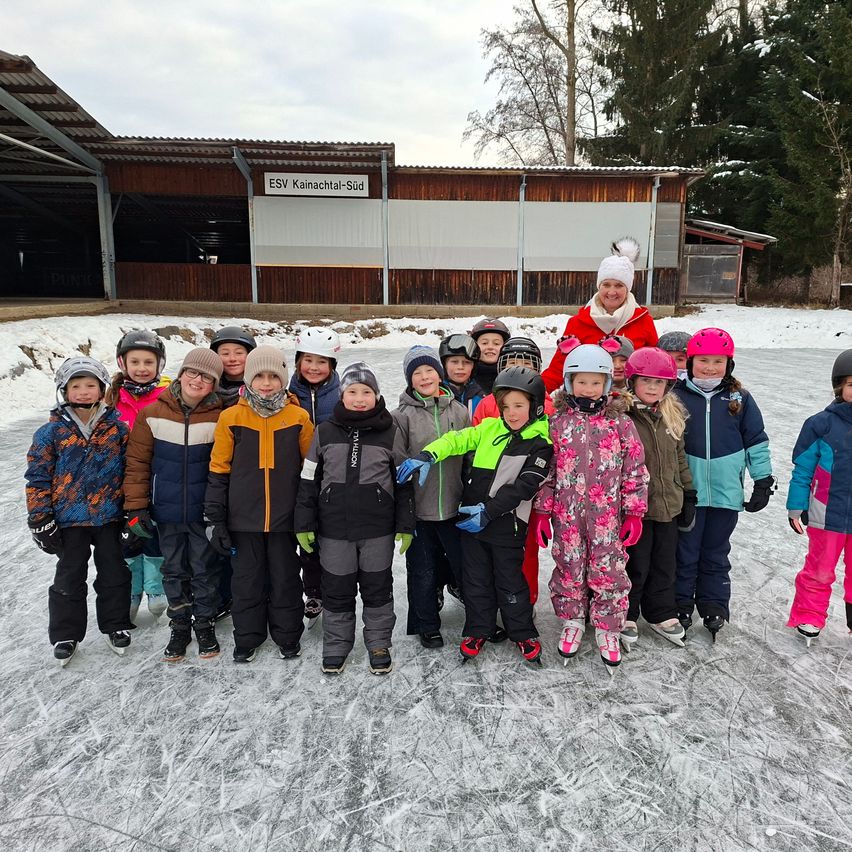 Eine Gruppe von Kindern und einer Frau in Winterkleidung posiert für ein Foto auf einer Eisbahn vor einem Gebäude.