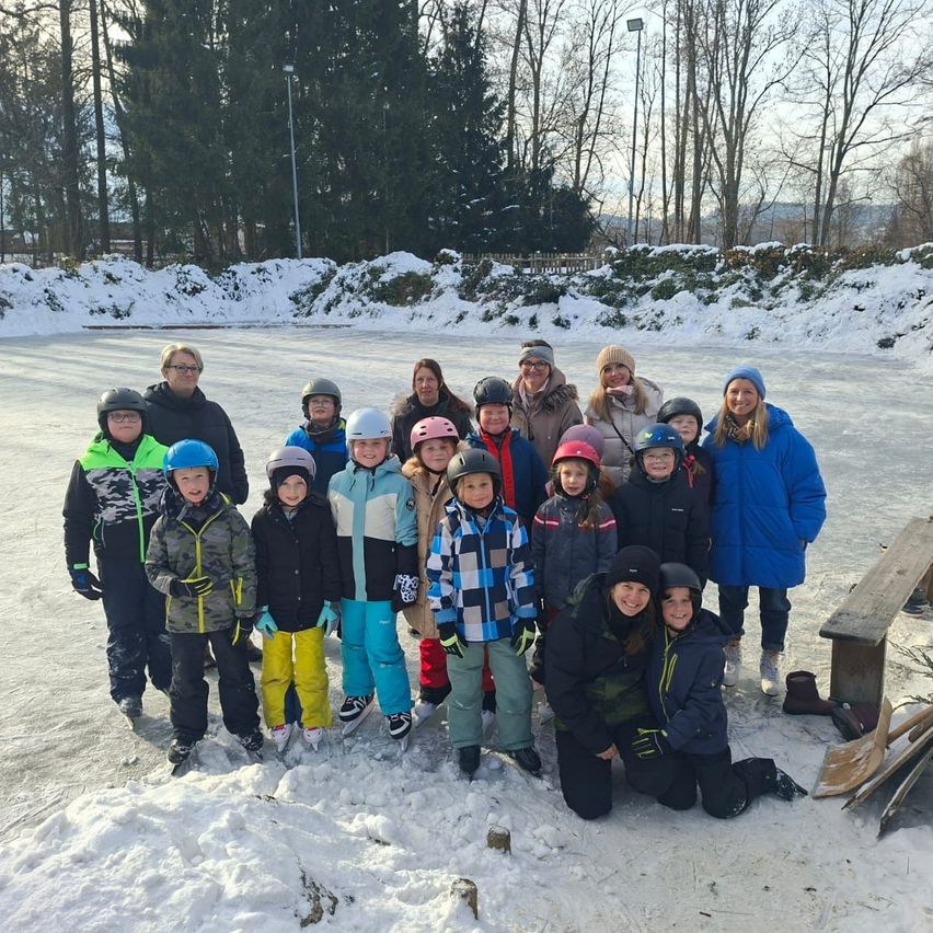 Eine Gruppe von Menschen, darunter Kinder, steht auf einem verschneiten Feld. Sie sind alle warm gekleidet und tragen Helme. Hinter ihnen ist eine Holzbank zu sehen.