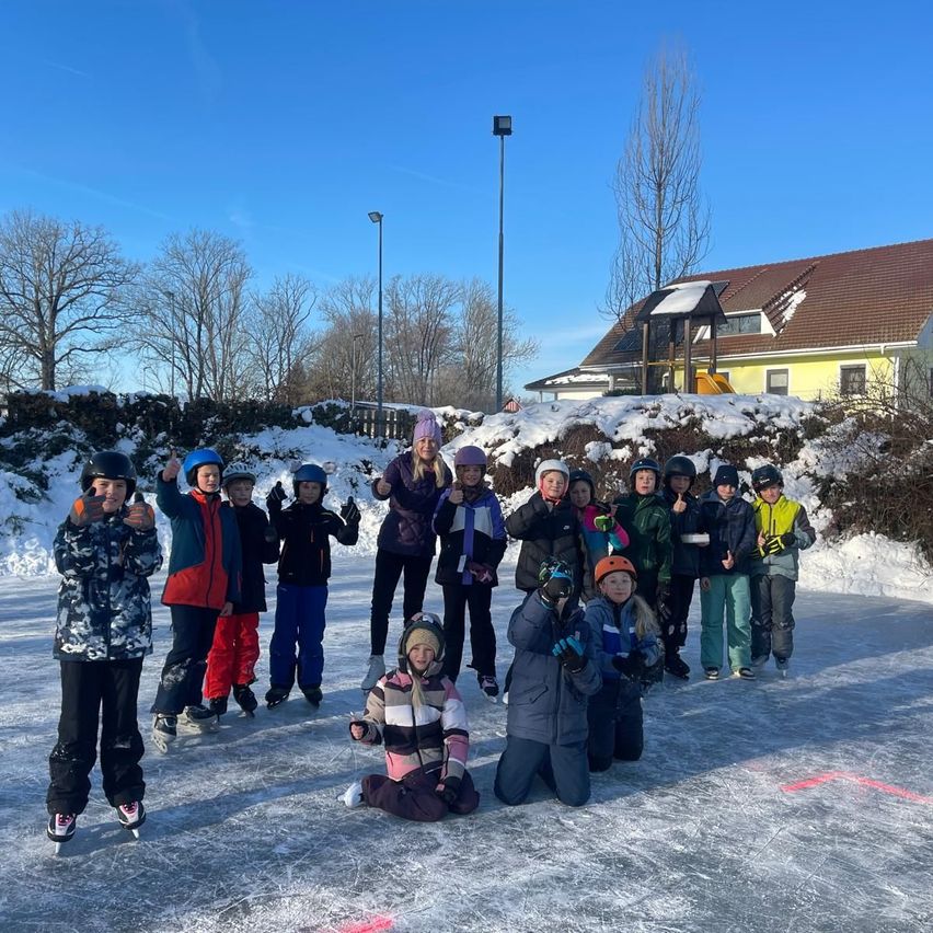 Eine Gruppe von Kindern und Erwachsenen posiert für ein Foto auf einer Eisbahn. Die Kinder tragen Winterkleidung und Helme. Hinter ihnen steht ein Haus mit einem braunen Dach und einer verschneiten Landschaft.