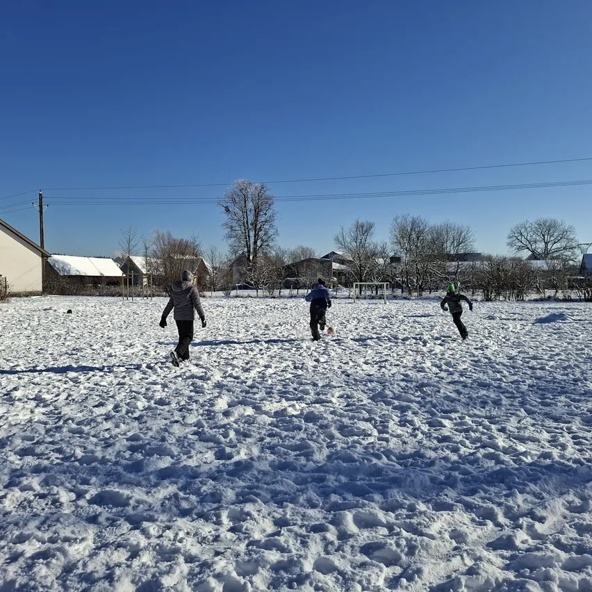 Drei Kinder rennen auf einem verschneiten Feld mit Häusern und Bäumen im Hintergrund unter einem klaren blauen Himmel.