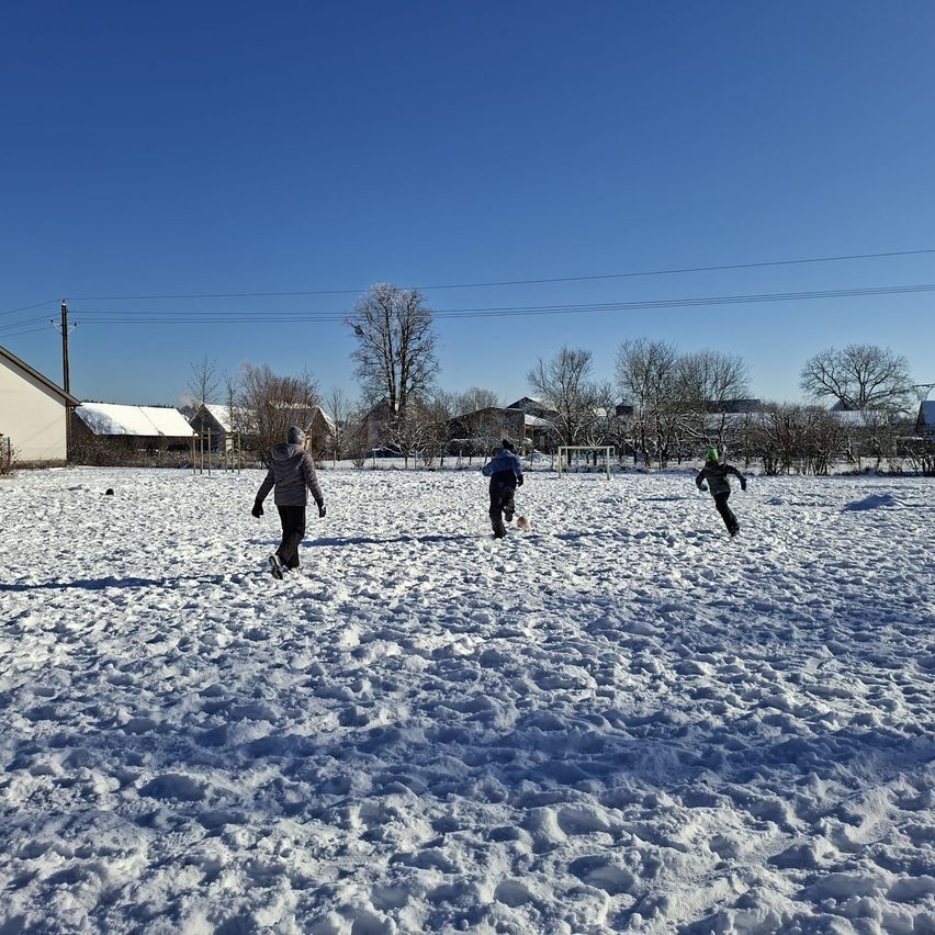 Drei Kinder rennen auf einem verschneiten Feld mit Häusern und Bäumen im Hintergrund unter einem klaren blauen Himmel.
