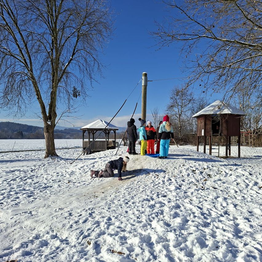 Mehrere Kinder in Winterkleidung spielen auf einem verschneiten Hang. Ein Kind rutscht hinunter, während andere zuschauen. In der Nähe stehen ein kleines Pavillon und eine Holzstruktur.