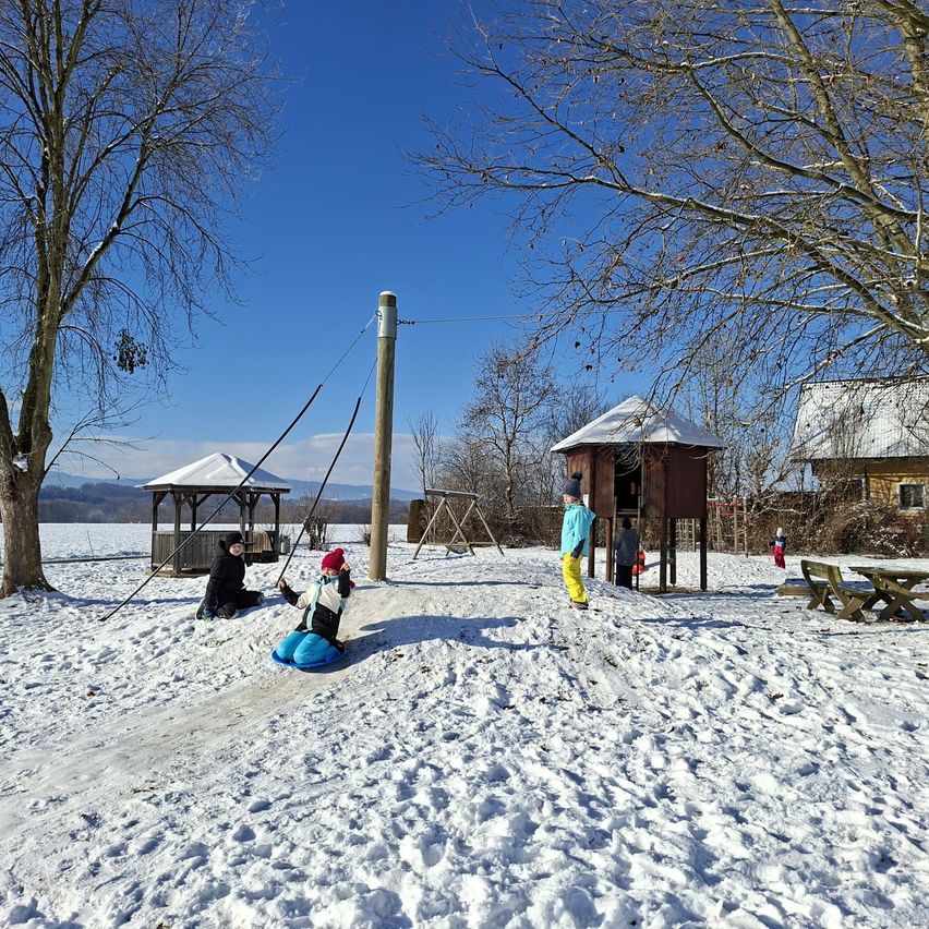 Ein Kind rutscht auf einem verschneiten Schlitten hinunter, während andere zusehen. Ein Pavillon und Picknicktische sind in der Nähe.