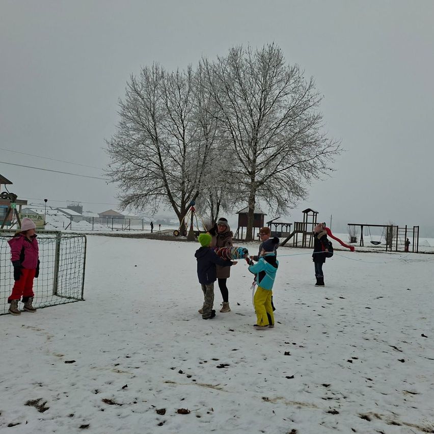Eine Gruppe von Kindern in Winterkleidung spielt im Schnee in der Nähe eines Spielplatzes. Sie führen einen Schneeballkampf, während eine Person ein Seil hält.
