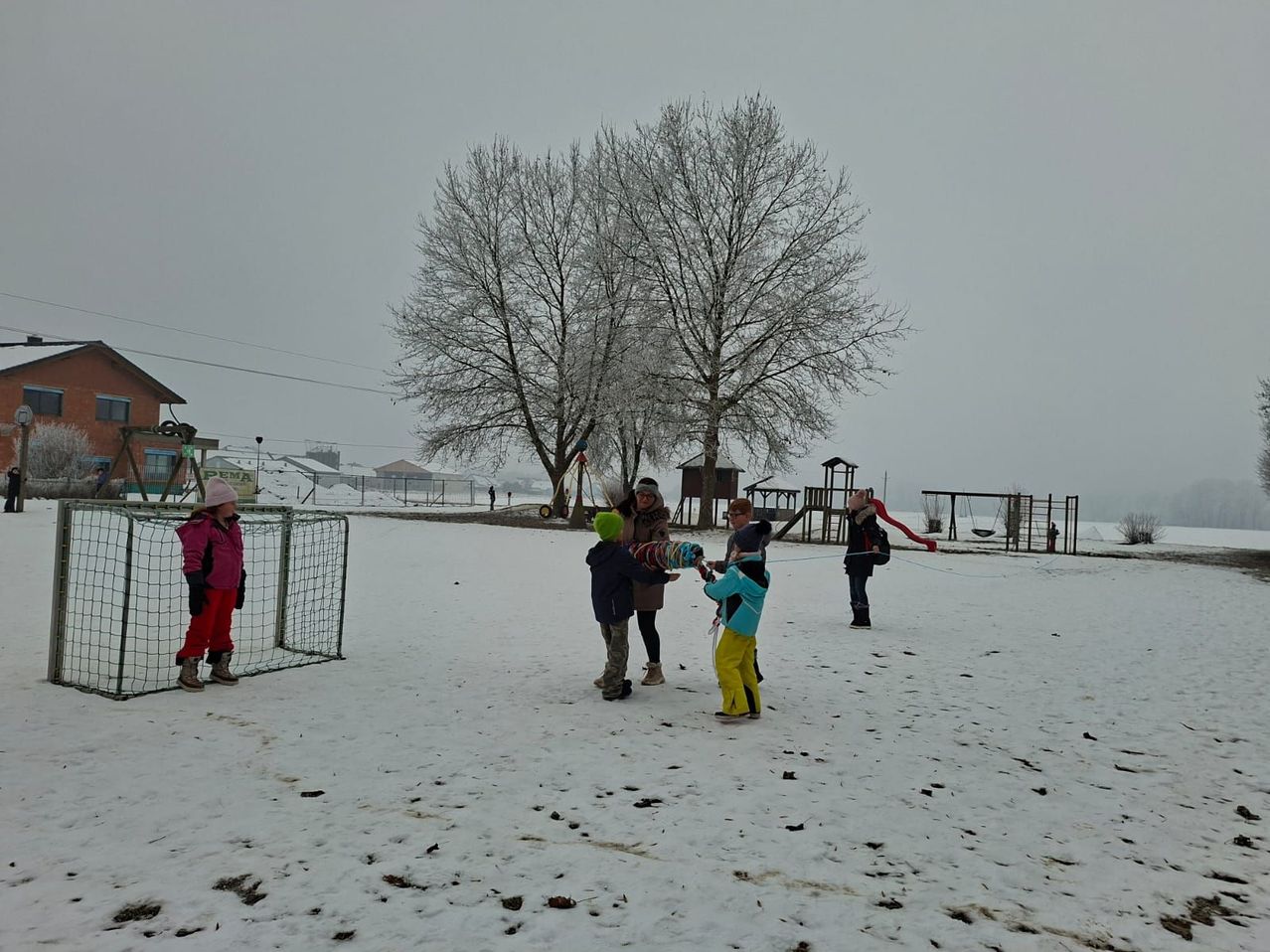 Eine Gruppe von Kindern in Winterkleidung spielt im Schnee in der Nähe eines Spielplatzes. Sie führen einen Schneeballkampf, während eine Person ein Seil hält.