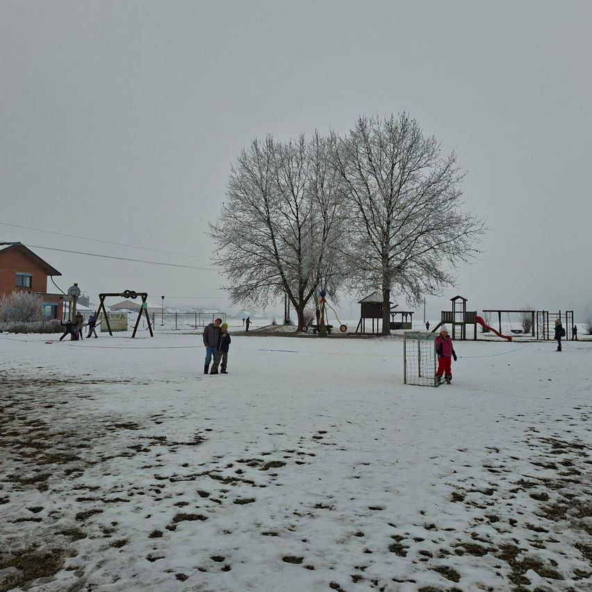 Ein schneebedecktes Feld mit Menschen und Bäumen. Ein Kind spielt auf der Rutsche. Ein Gebäude mit Glasfenstern befindet sich hinter dem Spielplatz.