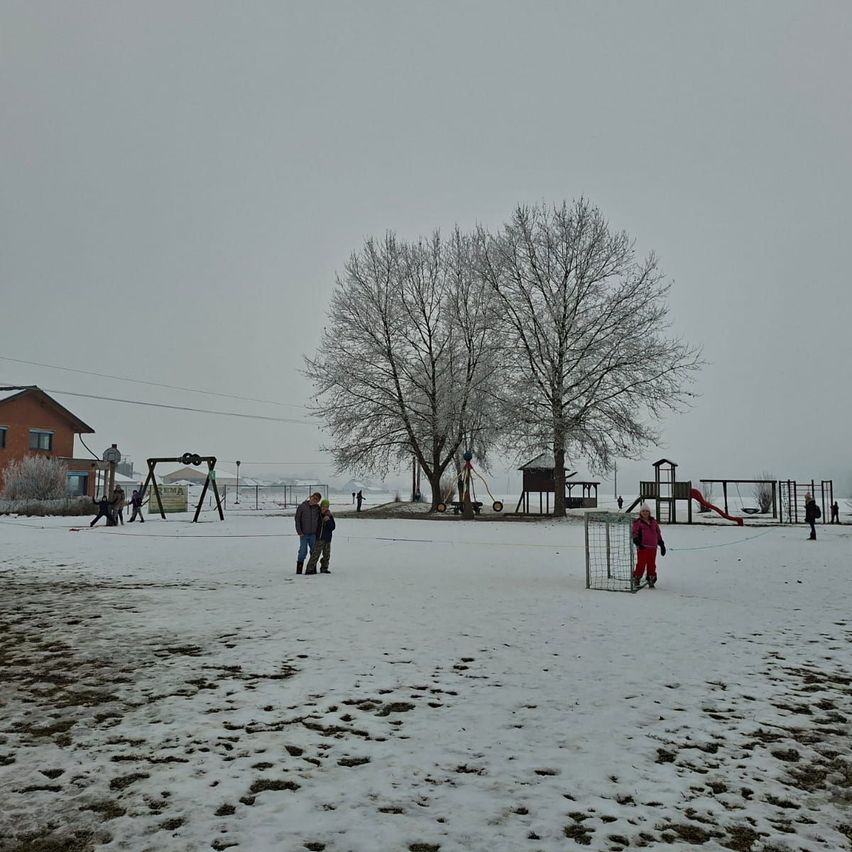 Ein schneebedeckter Spielplatz mit Menschen, die herumlaufen. In der Nähe steht eine Gruppe von Menschen neben einem Baum. Im Hintergrund befinden sich Rutschen und eine Schaukel.