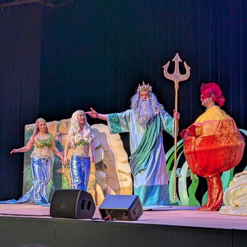 Auf der Bühne hält ein Mann in der Kleidung von Neptun einen Dreizack, flankiert von zwei Frauen in Meerjungfrauenkostümen. Ein dritter Mann in einer roten Kleidung steht in der Nähe. Schwarze Lautsprecher sind auf der Bühne positioniert.