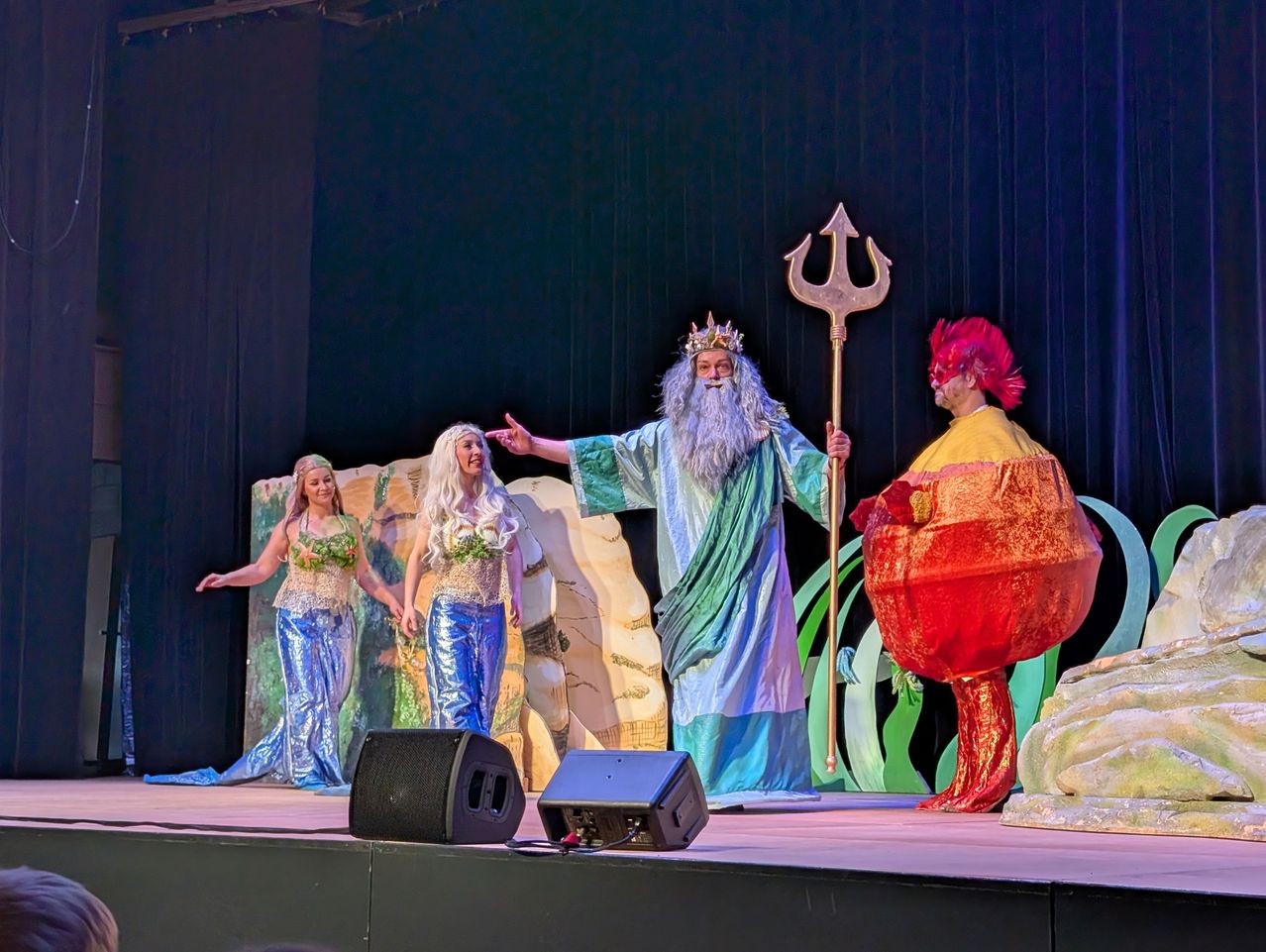 Auf der Bühne hält ein Mann in der Kleidung von Neptun einen Dreizack, flankiert von zwei Frauen in Meerjungfrauenkostümen. Ein dritter Mann in einer roten Kleidung steht in der Nähe. Schwarze Lautsprecher sind auf der Bühne positioniert.