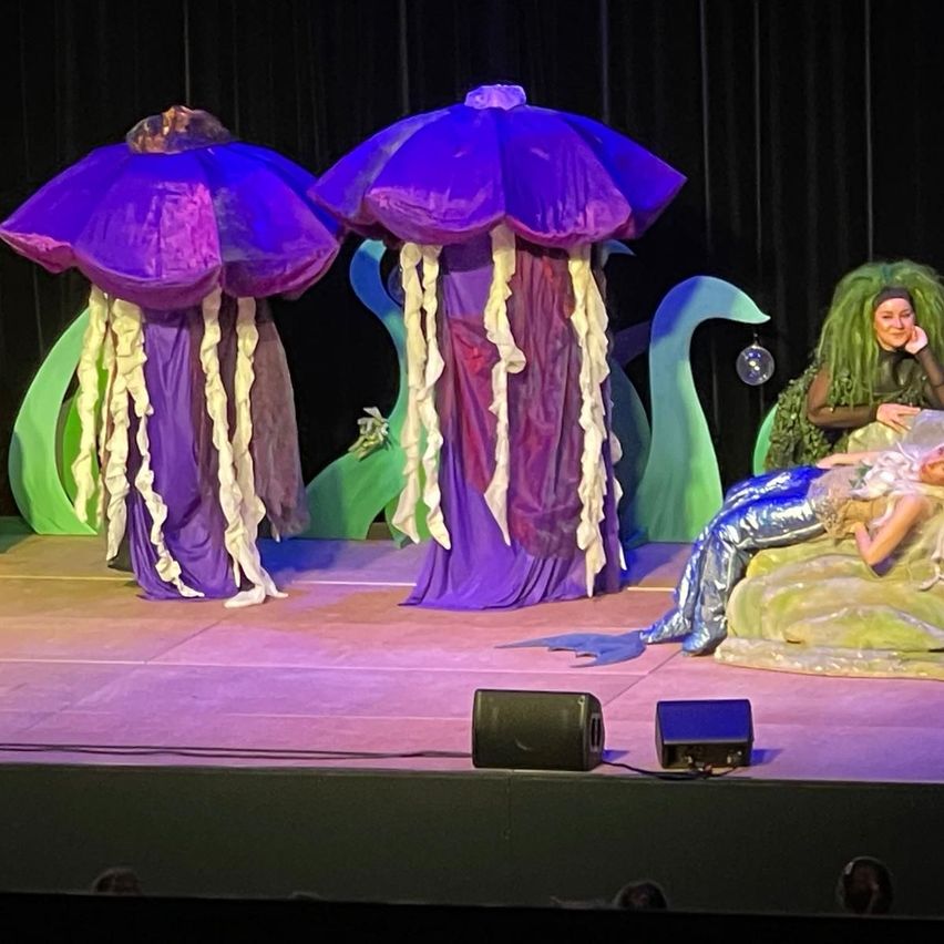 Auf der Bühne stehen zwei Darsteller in Kostümen von lila Meerestieren neben einer Frau in einem Meerjungfrauenkostüm, die auf einer grünen, algenartigen Struktur liegt. Die Bühne hat einen lila Boden und einen schwarzen Hintergrund.