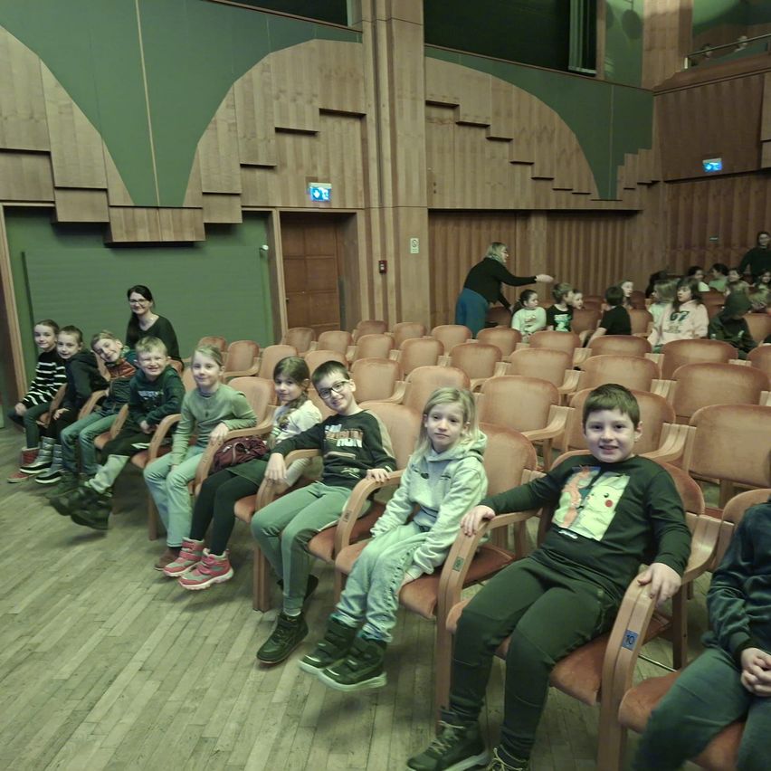 Eine Gruppe von Kindern sitzt in Reihen in einem Theater mit Erwachsenen, trägt Freizeitkleidung und Turnschuhe. Das Theater hat Holzsitze und Wände.