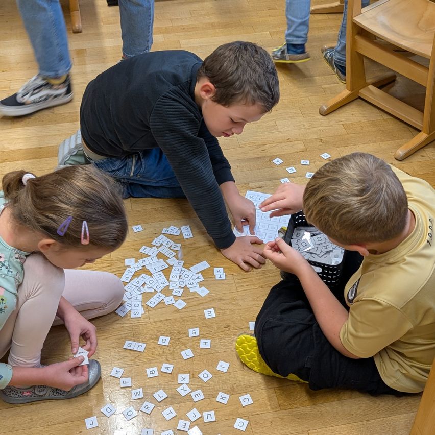 Drei Kinder spielen ein Wortspiel auf einem Holzboden, umgeben von verstreuten Buchstaben. Ein Junge in einem schwarzen Shirt greift nach Buchstaben, während ein Mädchen in einem Blumenshirt ein Stück hält. Ein anderer Junge in einem gelben Shirt schaut in einen Korb mit Buchstaben.