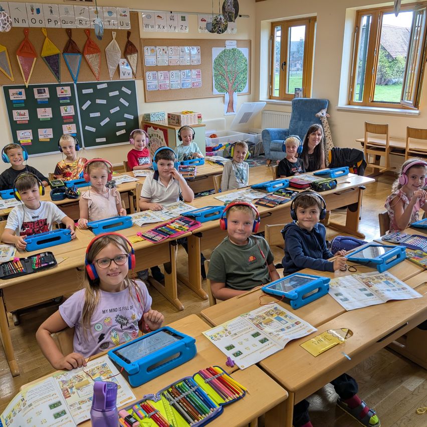 Ein Klassenzimmer mit jungen Schülern, die Kopfhörer tragen und an einer Aktivität mit Tablets und Malbüchern an ihren Schreibtischen teilnehmen. Eine Lehrerin ist anwesend, und es gibt Lernmaterialien an den Wänden.