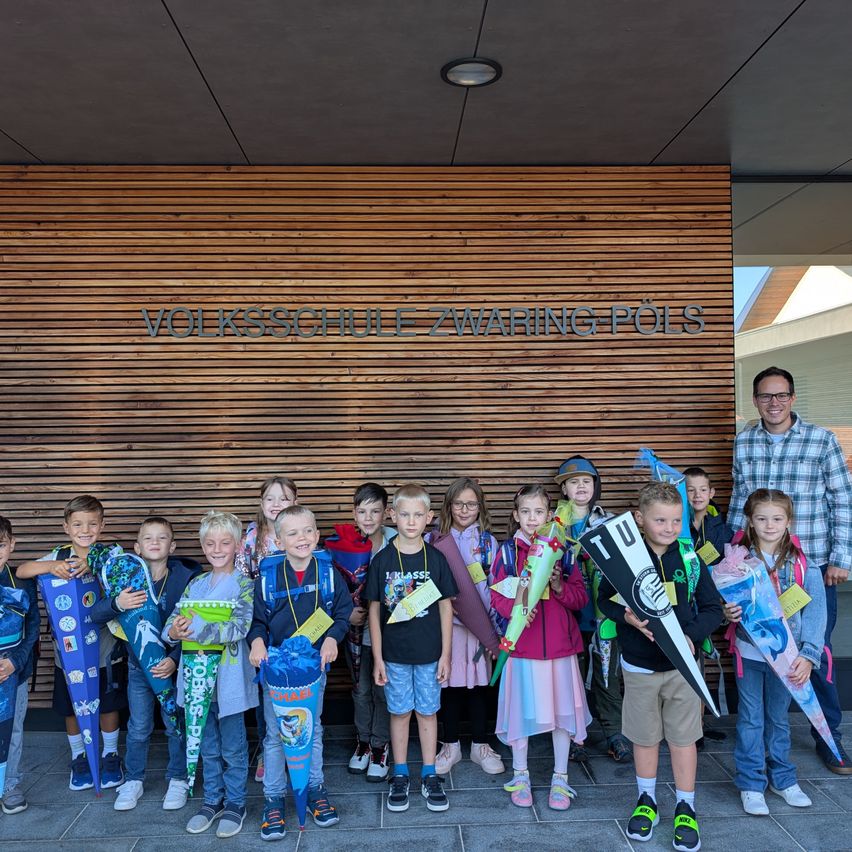 Eine Gruppe von Kindern, einige halten Blumen und andere Fahnen, stehen vor einem Gebäude mit dem Schild 'Volksschule ZWARING POLS'. Ein Mann steht neben ihnen und lächelt.