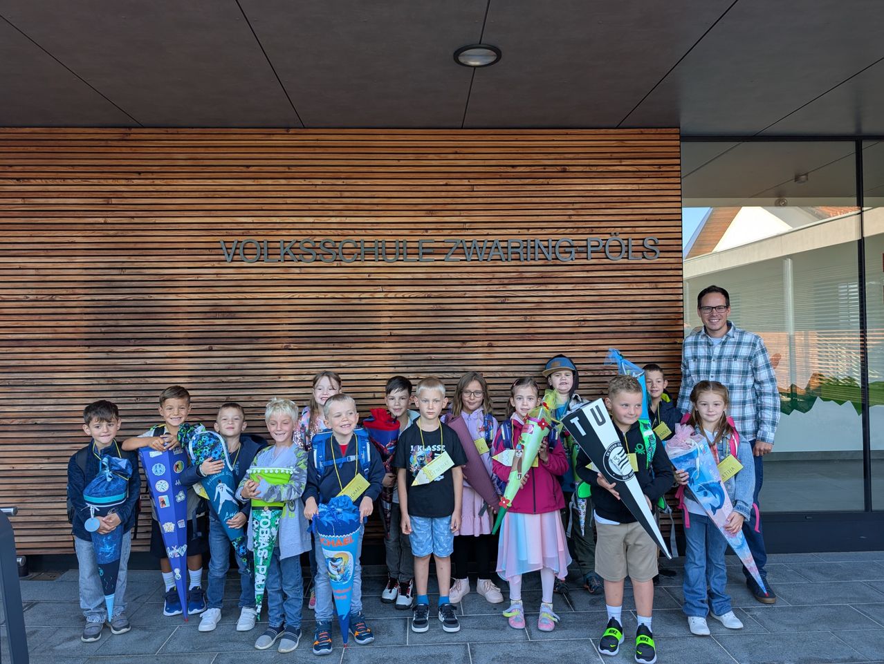 Eine Gruppe von Kindern, einige halten Blumen und andere Fahnen, stehen vor einem Gebäude mit dem Schild 'Volksschule ZWARING POLS'. Ein Mann steht neben ihnen und lächelt.