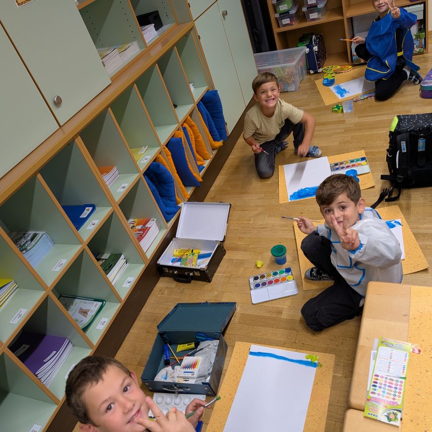 Eine Gruppe von Kindern sitzt auf dem Boden, lächelt und posiert für ein Foto. Sie sind umgeben von Malutensilien, einschließlich Pinsel, Paletten und Bechern. Der Raum ist gut beleuchtet, und es gibt Regale voller Bücher und anderer Gegenstände.