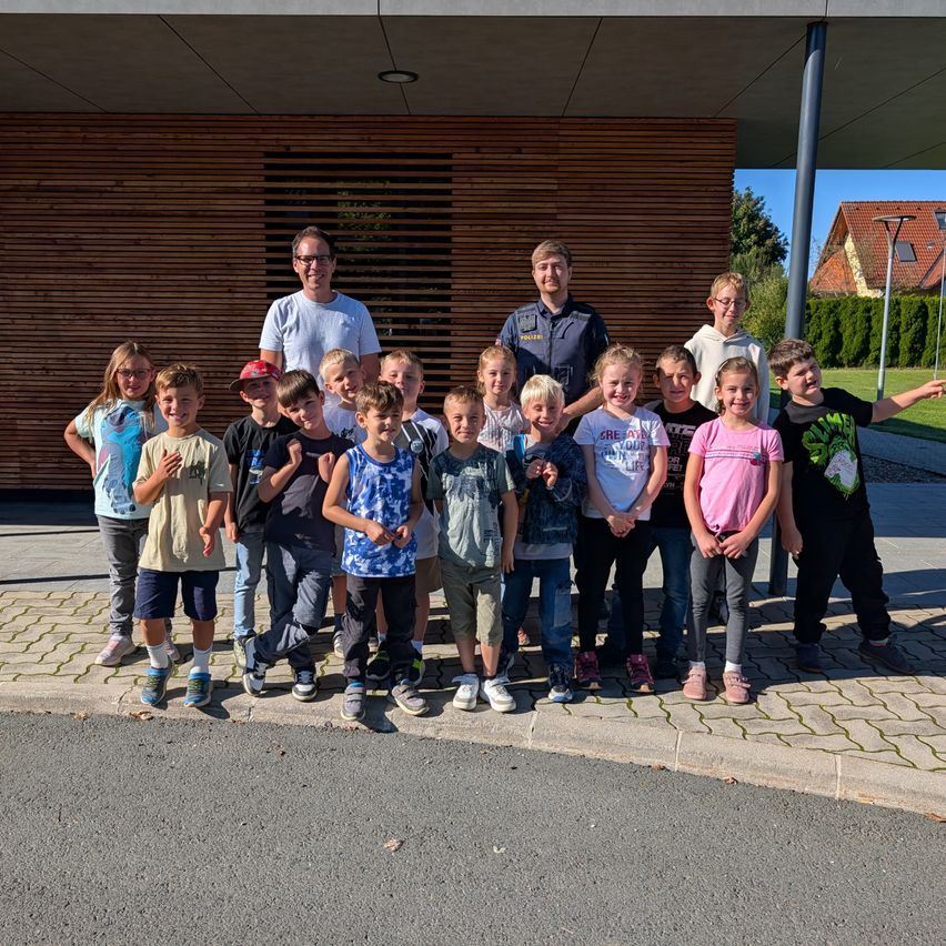 Eine Gruppe von Kindern und zwei Männern steht auf einem gepflasterten Bürgersteig vor einem Gebäude mit Holzwänden.