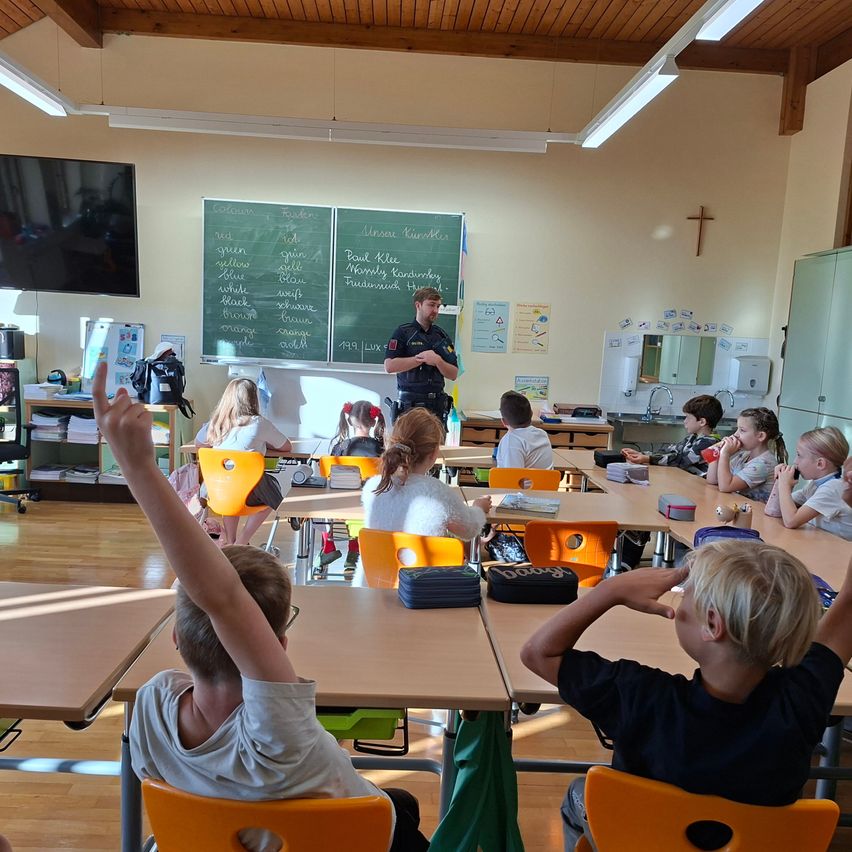 Ein Polizeibeamter unterrichtet eine Klasse in einem Klassenzimmer. Einige Schüler heben die Hände, andere schauen den Beamten an. Es gibt Tische, Stühle, eine Tafel und einen Fernseher.