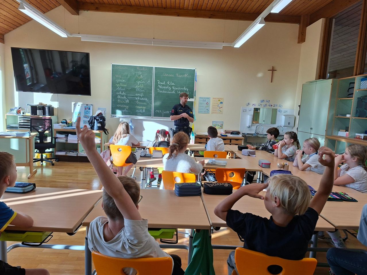 Ein Polizeibeamter unterrichtet eine Klasse in einem Klassenzimmer. Einige Schüler heben die Hände, andere schauen den Beamten an. Es gibt Tische, Stühle, eine Tafel und einen Fernseher.
