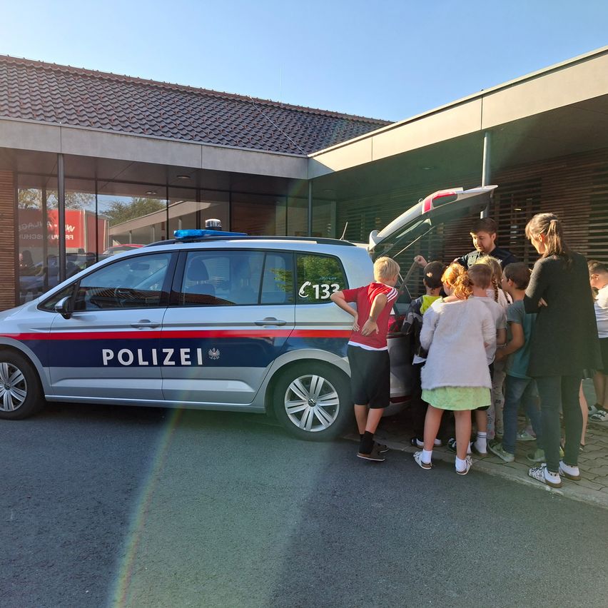 Mehrere Kinder und Erwachsene stehen um einen Polizeiwagen mit offenem Kofferraum herum. Das Auto hat die Nummer 133 und das Wort 'Polizei'. Ein Mann und eine Frau stehen mit den Kindern.