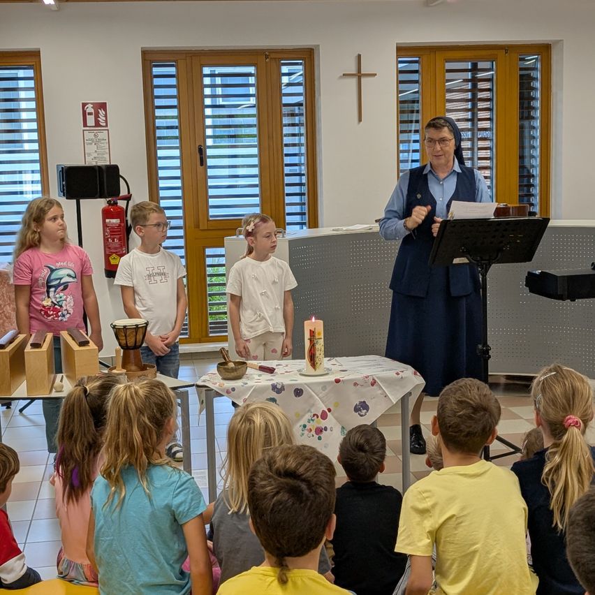 Eine erwachsene Frau spricht zu einer Gruppe von Kindern in einem Raum mit einem Kreuz an der Wand. Einige Kinder stehen hinter einem Tisch mit einer Kerze und einer Schüssel.