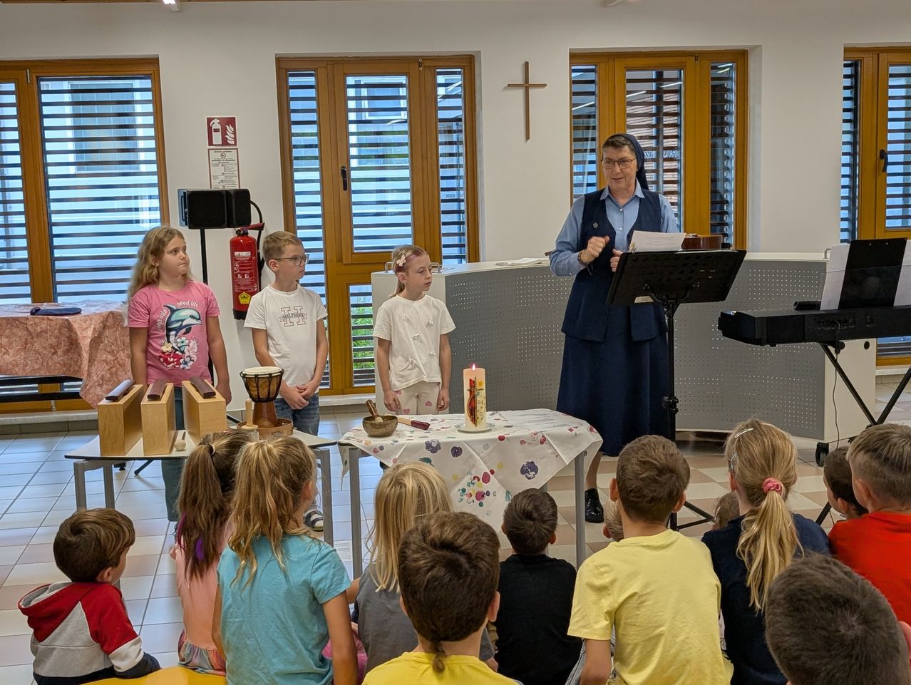 Eine erwachsene Frau spricht zu einer Gruppe von Kindern in einem Raum mit einem Kreuz an der Wand. Einige Kinder stehen hinter einem Tisch mit einer Kerze und einer Schüssel.