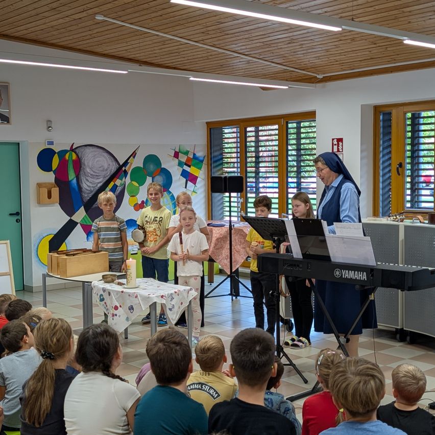 Eine Nonne steht vor einem Yamaha-Keyboard und unterrichtet eine Gruppe von Kindern in einem Klassenzimmer. Die Kinder halten Musikinstrumente in der Hand und stehen um einen Tisch mit einer Schüssel und einer Kerze herum.