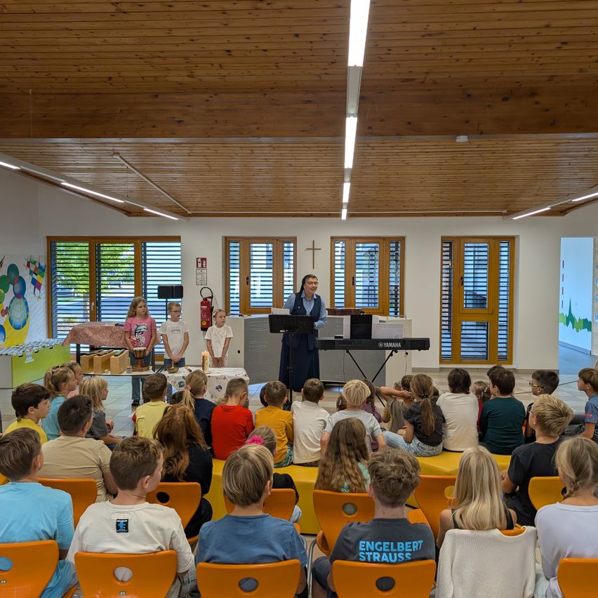 Eine Frau spricht an einem Podium in einem Raum mit Holzdecke und Wänden. Kinder sitzen in Stühlen vor ihr, und drei Kinder stehen neben einem Tisch.