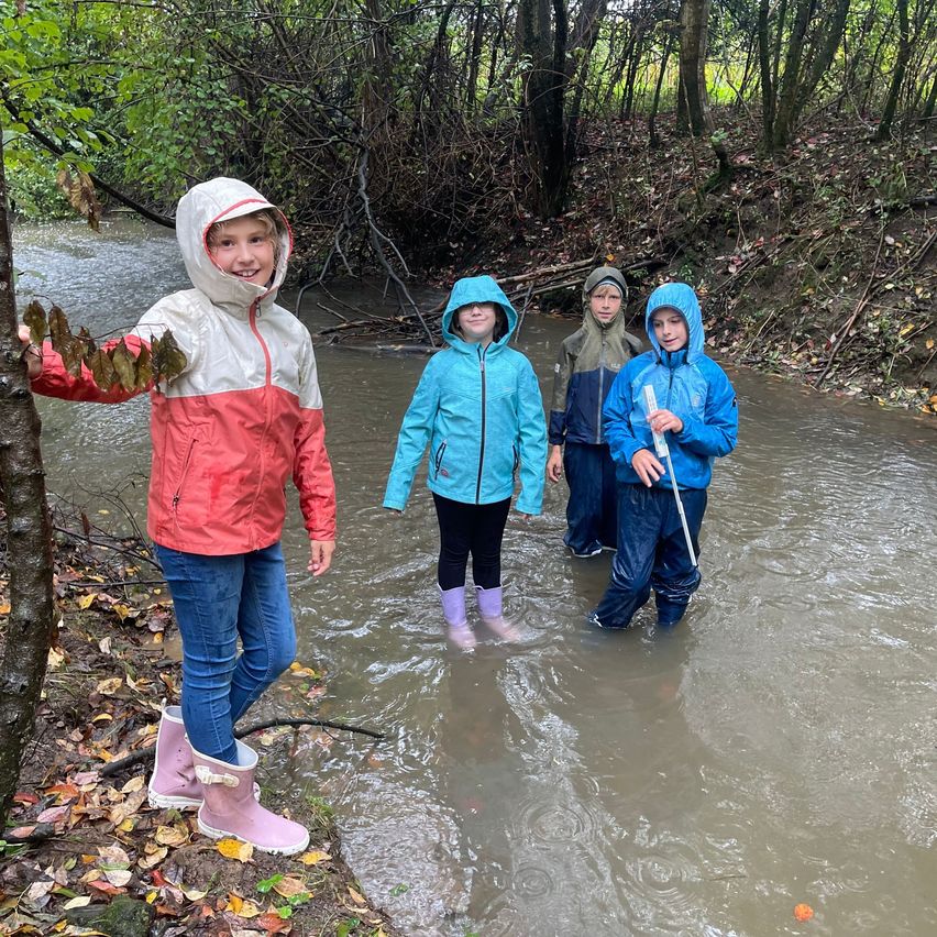 Vier Kinder stehen in einem Fluss, tragen Regenjacken und Stiefel. Sie lächeln, eines hält ein Blatt. Der Fluss ist von gefallenen Blättern und Bäumen umgeben.