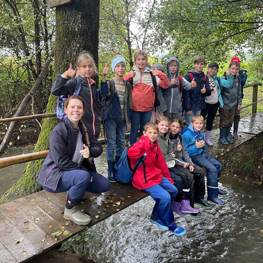 Eine Gruppe von Kindern, gekleidet in Jacken und Stiefeln, posiert für ein Foto auf einer Holzbrücke über einem Bach, umgeben von einem Wald.