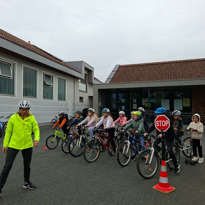 Eine Gruppe von Kindern mit Helmen fährt in einer Reihe auf Fahrrädern auf einem Parkplatz. Eine Frau steht davor und hält ein Stoppschild. Gebäude sind dahinter mit Fenstern und einem Schornstein.